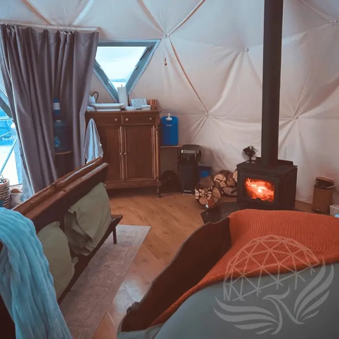 Cozy interior of the Worthwhile Escape geodesic dome in Alberta, featuring a crackling wood stove, rustic furnishings, and warm blankets for a comfortable glamping experience in all seasons.