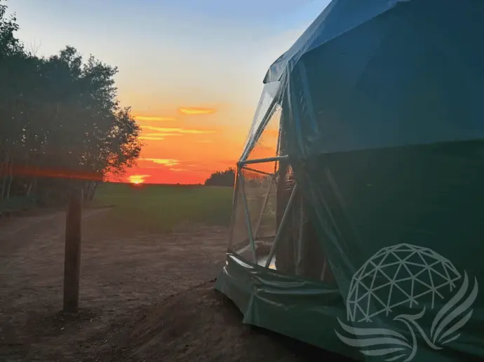 Sunset view from a glamping geodesic dome at Worthwhile Escape in Mayerthorpe, Alberta, with open farmland and forested trail visible under a glowing sky.