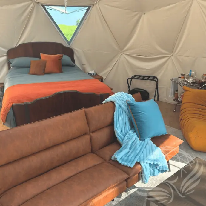 Interior of the geodesic glamping dome at Worthwhile Escape in Alberta, featuring a queen bed, cozy leather sofa, and vibrant throw blankets in a rustic-modern design.