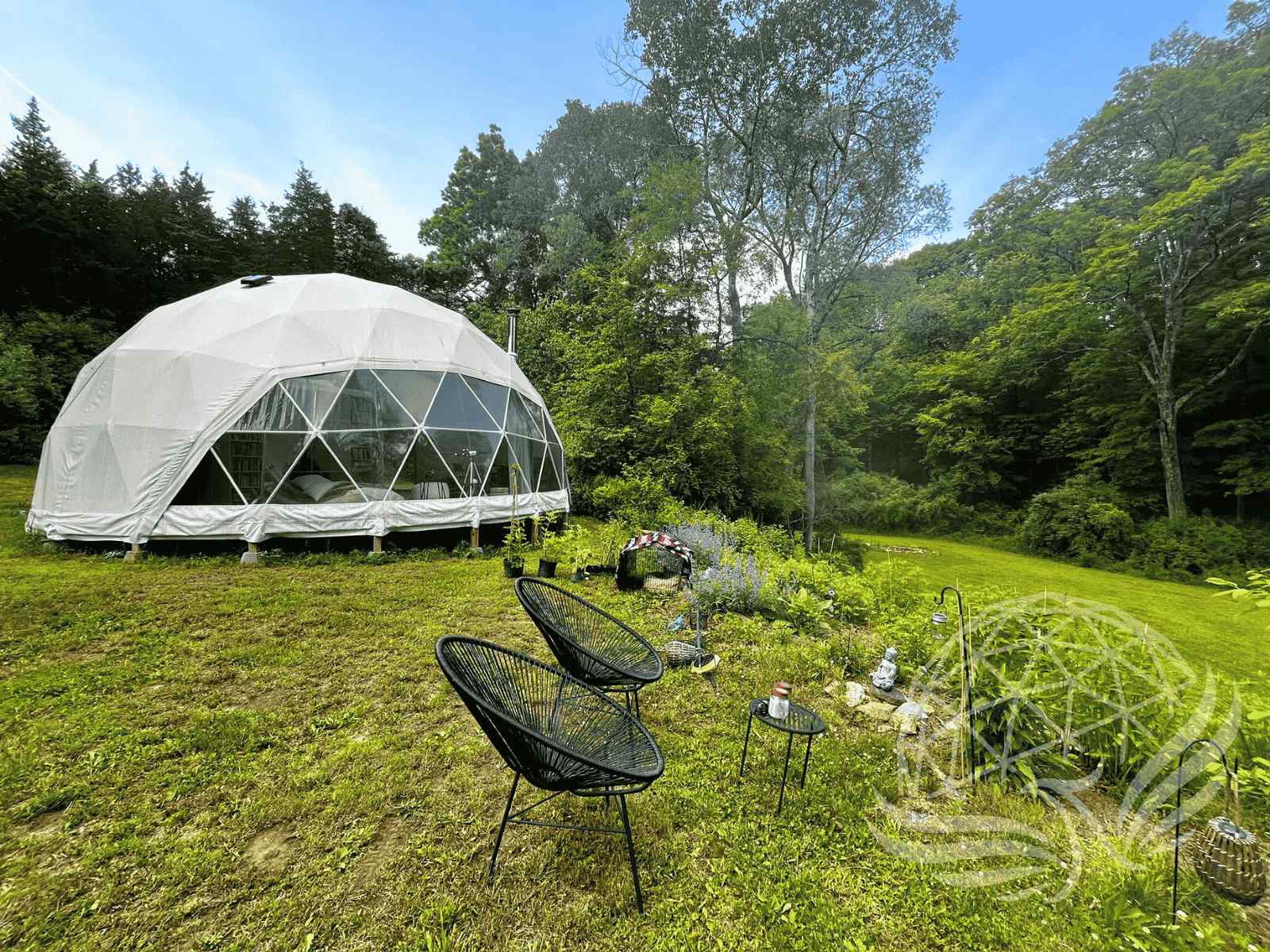 White geodesic dome with panoramic windows in a lush green meadow, surrounded by forest, with modern outdoor chairs and a small garden in the foreground.