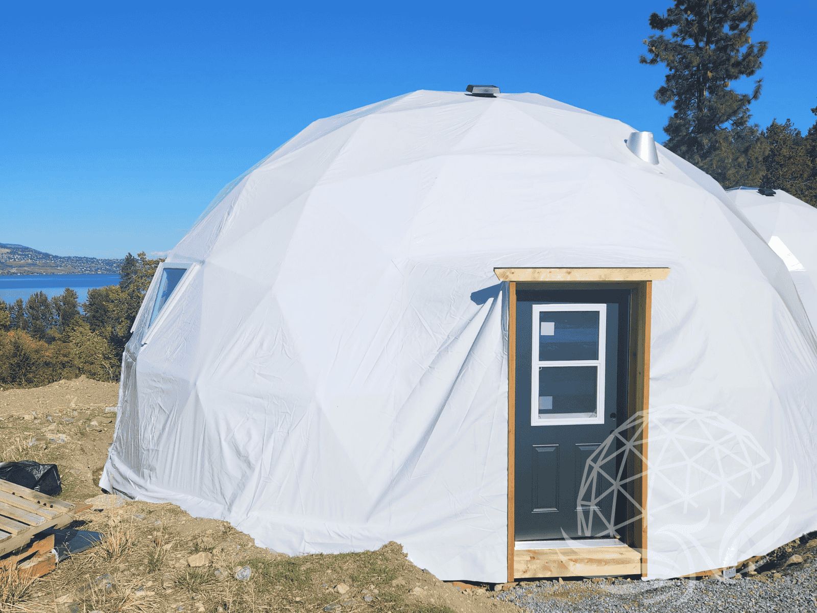 White geodesic dome with framed entry door on a hillside, overlooking a lake and forest with clear blue sky above.