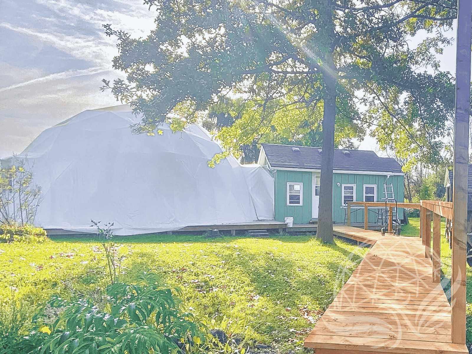 White geodesic dome beside a green cabin, surrounded by trees with a wooden boardwalk leading to the entrance, under warm sunlight.