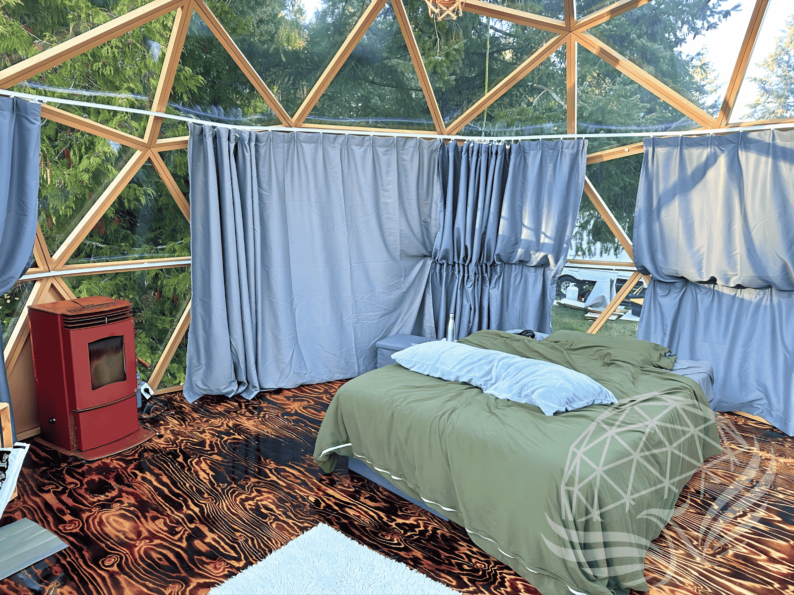 Cozy glass geodesic glamping dome by Phoenix Domes with bed, curtains, and stove on wood floor.